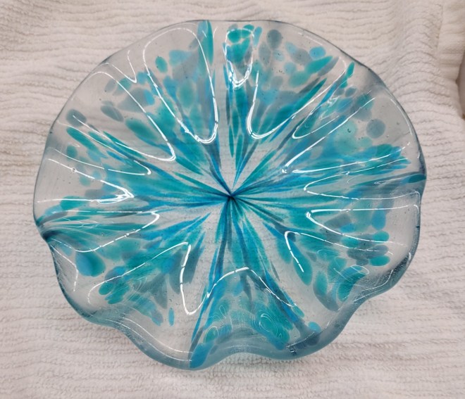 A wavy glass bowl made with a frit stretching technique. It is clear with varying shades of blue streaks that go to the center that look a bit like a firework.